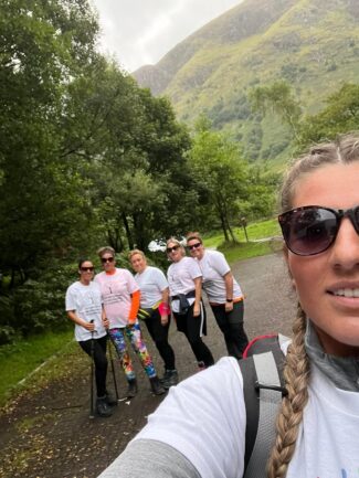A smiling woman in sunglasses in the foreground takes a selfie of herself and five others, in front of lush green countryside. The group is wearing Autism Centre of Excellence t-shirts and one has walking poles.