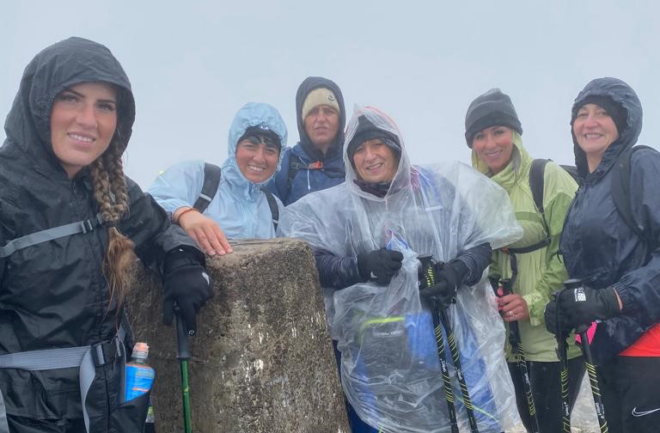 Six people in raincoats, with their hoods up, gather around a chest-height tree stump, smiling. The sky behind is grey and featureless.